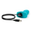 Na zdjęciu znajduje się turkusowy korek analny z czarnym kablem USB do ładowania, na którym widnieje logo "b-Vibe". Produkt ma zaokrąglony kształt i widoczny przycisk zasilania.