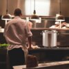 Na zdjęciu widać kuchnię restauracyjną z dużym garnkiem ze stali nierdzewnej na kuchence, na którym widnieje napis "KINGHOFF". W tle znajdują się kucharze przy pracy oraz wiszące lampy oświetlające przestrzeń roboczą.