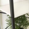 Zbliżenie na fragment regału z białą półką i czarną metalową ramą, z widoczną śrubą mocującą. W tle znajduje się zielona roślina o delikatnych liściach.