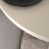 Okrągły stół o jasnej, gładkiej powierzchni z ciemnym talerzem na blacie, obok znajduje się szara tapicerowana powierzchnia. Podłoga ma wzór przypominający dywan w odcieniach szarości.