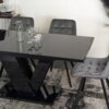 Czarny, połyskujący stół rozkładany z nowoczesnym designem, otoczony szarymi, tapicerowanymi krzesłami, stoi na wzorzystym dywanie. Na stole znajdują się szklana karafka i kilka kieliszków.