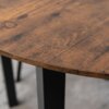 Zbliżenie na okrągły stół z blatem w kolorze dębu rustykalnego, ukazujący jego teksturę i naturalne słoje drewna. Nogi stołu są czarne i metalowe, widoczne w tle.