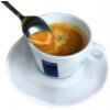 Biała filiżanka z logo "LAVAZZA" na spodku, wypełniona kawą espresso z kremową pianką, z łyżeczką unoszącą część pianki.