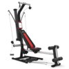 Na zdjęciu znajduje się atlas do ćwiczeń z czarno-czerwonym siedziskiem i oparciem, z widocznymi napisami "BOWFLEX" oraz logo producenta. Urządzenie posiada system linek i uchwytów do ćwiczeń siłowych.