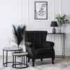 Czarny fotel wypoczynkowy z weluru stoi w elegancko urządzonym salonie, obok dwóch okrągłych stolików z przezroczystą karafką i szklankami. Na ścianie wisi minimalistyczny obraz, a na konsoli znajduje się lampa z ciemnym abażurem oraz wazon z zielonymi ga