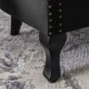 Czarny fotel wypoczynkowy z weluru, ozdobiony rzędem złotych nitów, stoi na dywanie o wzorze w odcieniach szarości. Widoczne są elegancko wyprofilowane, ciemne nogi fotela.