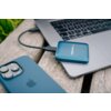 Na zdjęciu znajduje się niebieski dysk SSD marki Verbatim podłączony do laptopa, leżący na drewnianej powierzchni. Obok widoczny jest fragment smartfona w niebieskim etui.