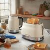 Na zdjęciu widać kremowy czajnik i toster marki Russell Hobbs na blacie kuchennym, obok znajdują się kromki tostów, słoiki z dżemem oraz filiżanka. W tle widoczna jest kuchnia z drewnianymi akcesoriami i rośliną w wazonie.