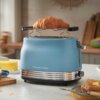 Niebieski toster marki Russell Hobbs z metalicznymi elementami, na którym znajduje się rogalik, stoi na blacie kuchennym. W tle widoczne są deski do krojenia, masło na talerzu oraz kawałki chleba na talerzyku.