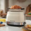 Na zdjęciu znajduje się jasny toster z logo "Russell Hobbs", na którym umieszczony jest rogalik. W tle widoczne są kuchenne akcesoria, takie jak deska do krojenia, masło i talerz z kromkami chleba.