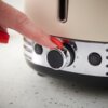 Zbliżenie na panel sterowania tostera z metalicznym pokrętłem, na którym widoczne są cyfry 5 i 6, oraz przyciskiem z symbolem płatka śniegu. Palec z czerwonym paznokciem dotyka pokrętła, a tło stanowi jasna, nakrapiana powierzchnia.