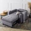 Szara, rozkładana sofa z podłokietnikami, na których znajdują się uchwyty na napoje, stoi na wzorzystym dywanie obok dwóch okrągłych stolików z książką i szklanym dzbankiem. Na sofie leży dekoracyjna poduszka w kształcie węzła oraz szary koc.