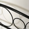 Czarne metalowe łóżko z ozdobnym wzorem z okręgów na wezgłowiu, widoczne na tle jasnej ściany. Konstrukcja jest gładka i lśniąca, z wyraźnymi cieniami rzucanymi na ścianę.