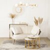 Kremowa sofa dwuosobowa w stylu glamour z poduszką i narzutą, ustawiona na jasnym dywanie z wzorem, przed dwoma okrągłymi stolikami z marmurowymi blatami i złotymi nogami. W tle znajduje się konsola z wazonem z suszonymi trawami, świecami i książkami, a n