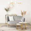 Na zdjęciu znajduje się szara sofa dwuosobowa w stylu glamour z białą i szarą poduszką, ustawiona na jasnym dywanie z wzorem. W tle widoczny jest złoty stolik z dekoracjami, okrągłe lustro z promienistą ramą oraz złoty żyrandol z białymi kloszami.