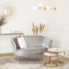 Szara sofa dwuosobowa w stylu glamour z dekoracyjnymi przeszyciami, obok której znajdują się dwa okrągłe stoliki z marmurowymi blatami i złotymi nogami. W tle widać lustro z ozdobną ramą, szklany stolik z książkami i świecami oraz dekoracyjne trawy w wazo