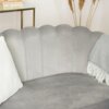 Szara sofa dwuosobowa o zaokrąglonym oparciu, z białą poduszką i szarym kocem z frędzlami. W tle widoczny jest szklany stolik z książkami oraz suszone trawy dekoracyjne.