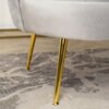 Szara sofa dwuosobowa w stylu glamour z widocznymi złotymi, metalowymi nogami stoi na dywanie o wzorze w odcieniach szarości. Tkanina sofy ma delikatną, aksamitną fakturę.