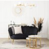 Czarna, dwuosobowa sofa w stylu glamour z dwoma poduszkami stoi na jasnym dywanie, obok niej znajdują się dwa okrągłe stoliki z marmurowymi blatami i złotymi nogami. W tle widać złote lustro w kształcie słońca, złoty żyrandol z białymi kloszami, konsolę z