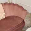 Różowa sofa dwuosobowa w stylu glamour z muszlowym oparciem, na której leży jasnoróżowy koc z frędzlami. W tle widoczna jest jasna ściana z listwą oraz fragment dywanu i stolika.