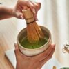 Na zdjęciu widoczne są dłonie trzymające ceramiczną miseczkę z zieloną herbatą matcha, w której zanurzone jest bambusowe mieszadło. Tło jest rozmyte, a na blacie leży metalowy przedmiot.