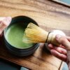 Na zdjęciu widać ceramiczną miseczkę z zieloną herbatą matcha oraz bambusowe mieszadło trzymane w dłoni. Tło stanowi drewniana powierzchnia.