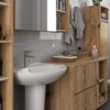 Na zdjęciu znajduje się zestaw mebli łazienkowych w kolorze dąb artisan i kaszmir, z widocznymi szafkami, półkami oraz umywalką. Na blacie stoi świeca, a na ścianie wiszą ręczniki i lustro.