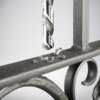 Wiertło metalowe znajduje się nad metalową powierzchnią, na której leżą wióry. W tle widoczna jest część metalowej konstrukcji z ozdobnym elementem.