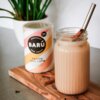 Puszka z napisem "BARÚ Vanilla Chai Latte" stoi na drewnianej desce obok szklanki z napojem, w której znajduje się metalowa słomka i laska cynamonu. W tle widoczna jest zielona roślina w białej doniczce.