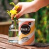 Puszka z napisem "BARÚ Vanilla Chai Latte with black tea & exotic spices" oraz "VEGAN RECIPE" stoi na drewnianym stole obok szklanej butelki z wodą i żółtymi kwiatami, które są trzymane przez rękę. W tle widoczna jest rozmyta zieleń.
