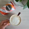 Puszka z napisem "BARÚ Spiced Chai Latte Powder with Real Spices" leży na drewnianym blacie obok filiżanki z napojem o kremowej powierzchni, trzymanej przez dłoń z pomalowanymi na brązowo paznokciami. W tle widoczna jest biała taca z zieloną rośliną.