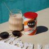 Na zdjęciu znajduje się puszka napoju "BARÚ Spiced Chai Latte" z czerwonym wieczkiem, obok szklanka z napojem i słomką, a także okulary przeciwsłoneczne leżące na białym ręczniku z frędzlami. W tle widoczny jest basen.