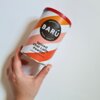 Puszka z napisem "BARÚ Surreally Good Treats Spiced Chai Latte Powder with Real Spices" trzymana w dłoni, ma kolorowe, faliste wzory i czerwone wieczko.