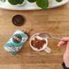 Na drewnianym blacie znajduje się otwarte opakowanie z napisem "BARÚ Fluffy Marshmallow Chocolate Powder" oraz filiżanka z napojem posypanym czekoladowym proszkiem, obok której trzymana jest łyżeczka. W tle widoczna jest biała taca z zielonymi roślinami.