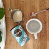 Na drewnianym blacie znajduje się puszka z napisem "BARÚ Fluffy Marshmallow Chocolate Powder", obok której stoi metalowy dzbanek z mlekiem, filiżanka oraz łyżeczka. W tle widoczny jest fragment zielonej rośliny.