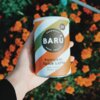 Puszka z napisem "BARÚ Surreally Good Treats Pumpkin Spice Latte with coffee & exotic spices" trzymana w dłoni, na tle kwitnących pomarańczowych kwiatów. Na opakowaniu widnieje również oznaczenie "Vegan Recipe".