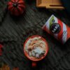 Na zdjęciu znajduje się pomarańczowy kubek z napojem pokrytym bitą śmietaną, obok leży opakowanie z napisem "BARÚ Pumpkin Spice Latte with coffee & exotic spices". W tle widoczna jest mała, dekoracyjna dynia oraz kolorowe liście na ciemnym, teksturowanym materiale.