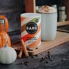 Na zdjęciu znajduje się opakowanie napoju "BARÚ Pumpkin Spice Latte" z napisem "Surreally Good Treats" oraz "Vegan Recipe", umieszczone na drewnianej desce obok filiżanki z pianką, lasek cynamonu, ziaren kawy i małych dyń. Tło stanowi ciemny blat i rozmyte elementy kuchenne.