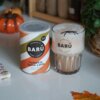 Puszka z napisem "BARÚ Pumpkin Spice Latte with coffee & exotic spices" stoi obok szklanki z napojem, na której widnieje logo "BARÚ". W tle widoczna jest dekoracja w postaci dyni oraz zielona roślina.
