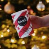 Puszka z napisem "BARÚ Swirly Hot Chocolate Powder" trzymana w dłoni na tle rozmytych światełek choinkowych. Na opakowaniu widnieje również napis "Vegan Recipe" oraz "with chocolate figurine inclusions".