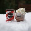 Puszka z napisem "BARÚ Swirly Hot Chocolate Powder" stoi na śniegu obok szklanki z gorącą czekoladą, na której widać bitą śmietanę. Na puszce i szklance widnieje logo "BARÚ Surreally Good Treats".