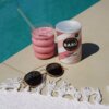 Na zdjęciu znajduje się puszka z napisem "BARÚ Pink Chai Latte" oraz szklanka z różowym napojem i szklana słomka, ustawione na brzegu basenu. Obok leżą okulary przeciwsłoneczne na białym, frędzlowanym ręczniku.