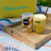 Na zdjęciu znajduje się puszka z napisem "BARÚ Golden Chai Latte" oraz szklanka z napojem o żółtym kolorze, ustawione na drewnianej tacy. W tle widoczny jest napis "Polarbox" na żółtym tle.