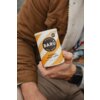 Osoba trzyma w ręku opakowanie napoju "BARÚ Golden Chai Latte" o pojemności 250 g, z widocznym napisem "Unreally Good Treats" oraz informacją "Vegan Recipe". Opakowanie ma żółto-białe paski i znajduje się na tle brązowej kurtki z futrzanym kołnierzem.