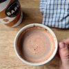 Na zdjęciu widoczny jest kubek z napojem czekoladowym, trzymany w dłoni, obok znajduje się puszka z napisem "BARÚ Salty Caramel Hot Chocolate" oraz ściereczka w niebieską kratkę. Tło stanowi drewniany blat.