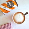 Puszka z napisem "BARÚ Surreally Good Treats Dark Hot Chocolate Powder 64% Cacao" leży obok filiżanki z napojem o kremowej piance. Na dole zdjęcia widoczna jest czerwona ściereczka w białą kratkę.