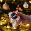 Puszka z napisem "BARÚ Dark Hot Chocolate Powder 64% Cacao" trzymana w dłoni na tle rozmytych światełek choinkowych. Opakowanie ma pomarańczowo-białe paski i czarne wieczko.