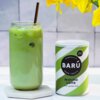 Na zdjęciu znajduje się szklanka z zielonym napojem z lodem i metalową słomką oraz puszka z napisem "BARÚ Surreally Good Treats Matcha Latte with green tea" i oznaczeniem "Vegan Recipe". W tle widoczne są żółte kwiaty.