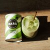Puszka z napisem "BARÚ Matcha Latte with green tea" stoi obok szklanki z zielonym napojem i kostkami lodu, w której znajduje się metalowa słomka. Na puszce widnieje również napis "VEGAN RECIPE".