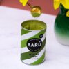 Puszka z napisem "BARÚ Surreally Good Treats Matcha Latte with green tea" oraz "Vegan Recipe" stoi na blacie, a nad nią unosi się łyżeczka z sypkim, zielonym proszkiem. W tle widoczny jest fragment zielonego wazonu z żółtymi kwiatami.